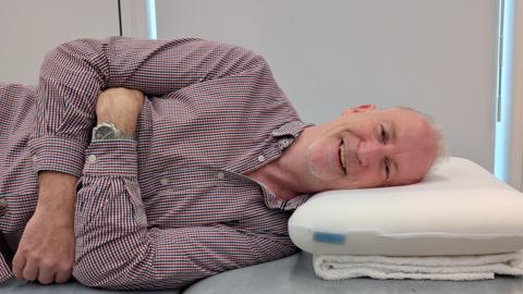 Person lying on their side on a padded treatment table with their head resting on a foam pillow. The person is wearing a checked button-up shirt and a wristwatch, with arms folded across the torso. The pillow is positioned on a folded towel. The window blinds in the background are drawn.