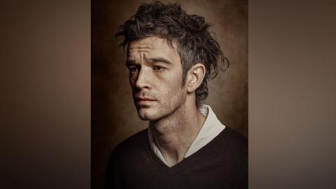 The portrait of Matty Healy which has him looking to the left with a serious look on his face and slightly furrowed brow. He is sitting in front of a grainy, brown backdrop which has creates a realist, gritty effect. Healy has dark hair which is greying on the sides and is cut in a mullet where it is longer on the top and shaved short on the sides. He has dark stubble around his mouth and down his neck. He is wearing a dark v-neck jumper with a white shirt underneath.