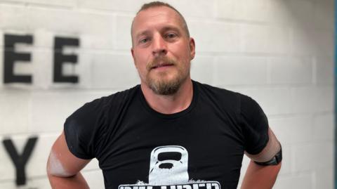 A man with short hair and a short beard and moustache is looking at directly at the camera. He wears a black t-shirt with an outline of a kettlebell on it