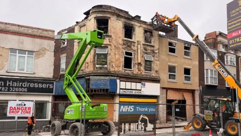 A building that has been ravaged by fire. There are two telehandlers extended out with workmen visible on one inspecting the damaged building.