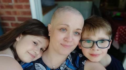 A woman who has lost her hair is being hugged by two children. They appear to be standing outside a house.