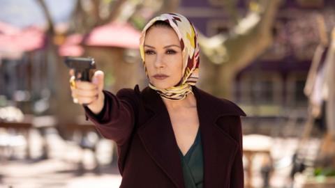 Catherine Zeta-Jones holding a gun up, wearing a scarf around her head and a maroon jacket in the Kill Jackie series.