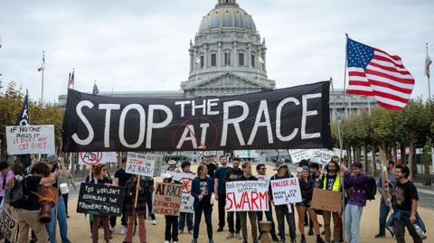 Protesters at an AI demonstration outside City Hall in San Francisco, California. Some in the crowd are holding a large black "Stop the AI race" banner, while others hold placards calling for a pause to AI development.