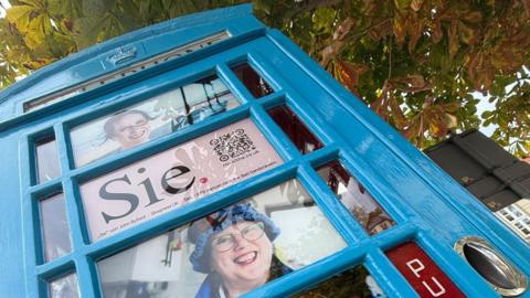 A blue telephone box is photographed from a low angle beneath the branches of a tree. Each window is filled with a portrait of a woman. One is filled the word “Sie,” and a QR code. 
