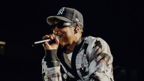 Jay-Z raps into a microphone while performing on stage. He wears a black and white cardigan and a New York Yankees hat.