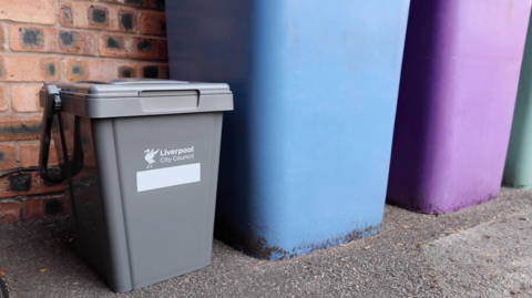 A small grey plastic bin with Liverpool City Council written on it stands next to taller blue and purple bins