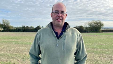 James Winslade is standing in a field and is wearing a green jumper. He is wearing glasses and is looking at the camera. 