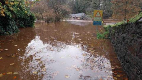 Severe flooding at Jubilee Bridge in Appleby. There is a road sign reading Ford, road liable to flooding. The water is a muddy brown colour with fallen leaves floating on the surface. A footbridge is in the distance with the bottom of its staircase submerged.