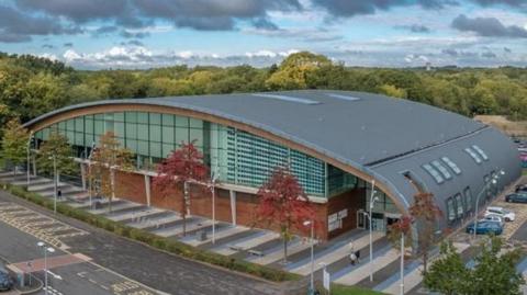 The outside of Corby East Midlands International Pool, showing a large building, with a curved roof, with a row of trees outside it, and trees behind it. The roof is grey, with windows in it, and a red bricks at the front with large glass windows at the front. Cars are parked to the right.