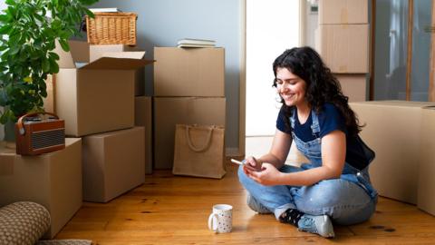 A youn woman sits on teh floor in her new home surrounded by moving in boxes
