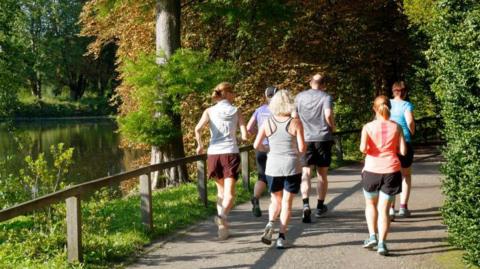 A group of people going for a run in parkland. There is a body of water to the left of the image, with trees and a small wooden fence