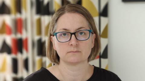 Esther Taylor is pictured in a head and shoulders image which shows her looking very serious. She has blonde hair which is in a 'bob' hairstyle and she is wearing a black top and blue glasses. Patterned curtains can be seen behind her.