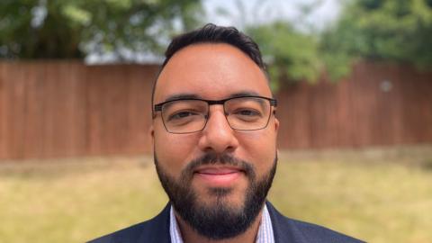 Man wearing a blue suit and checked shirt looks at the camera. He is wearing black glasses and has black hair and a beard.