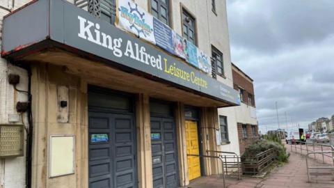 The entrance to the King Alfred Leisure Centre in Hove. A large sign above the entrance reads "King Alfred Leisure Centre", with banners promoting activities at the facility. The frontage shows ageing concrete, dark entrance doors and a ramp leading to the building. Kingsway runs alongside the leisure centre, with traffic and seafront buildings visible in the background.