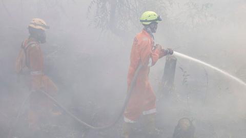 Two firefighters in bright orange uniform and helmets, standing outdoors in thick white haze, spray water from a hose