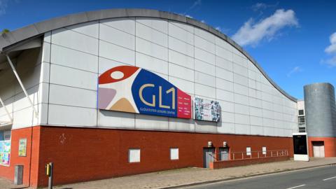 The side of GL1 building with a road in front of it. The sky behind is blue with clouds.