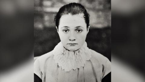 A black and white photograph of a young woman with hair tied back, looking straight at the viewer. She is wearing a white, frilly ruffle over a light-coloured top.
