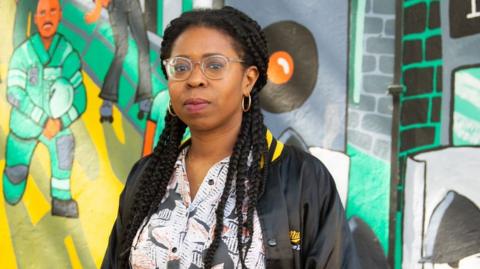 Stephanie Phillips has long braided hair. She is wearing glasses and hooped earrings with a black and white printed dress with a black bomber jacket over. She is standing in front of a colourful mural painted on the wall. 
