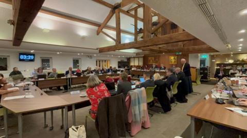 A photograph of the council chambers. There's dozens of people sitting around multiple tables arranged in a rectangle. Above those sat around in the council chamber on green chairs, there's high wooden beams along with a wooden back to the room.