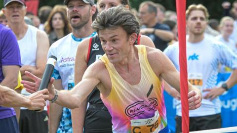 A runner being passed a baton during a relay race. He is wearing a yellow and purple vest with the word "FLYCARB" printed on the front.