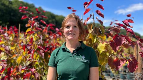 Stephanie James is wearing a green polo shirt with the nursery's logo on. She has brown hair tied loosely back and brown eyes. She is smiling. Behind her are rows of trees in pots, they are cercis eternal flame, the leaves are a vibrant green, yellow and red.