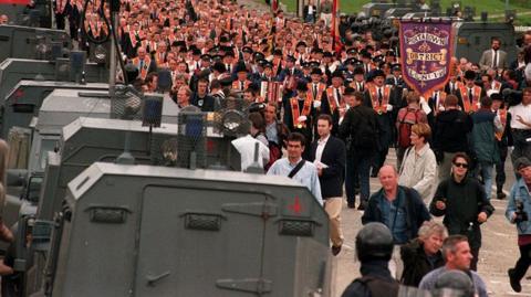 A large parade is on a road. Men are wearing orange sashes and black bowler hats. Police range rovers are parked on the road. Some men are holding banners and flags. 
