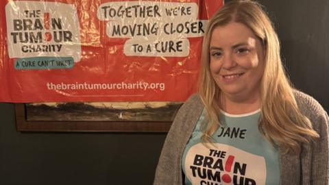 Jane is wearing a Brain Tumour Charity T-shirt and is stood in front of a similar banner on a wall behind her. She has long blonde hair and is smiling at the camera.