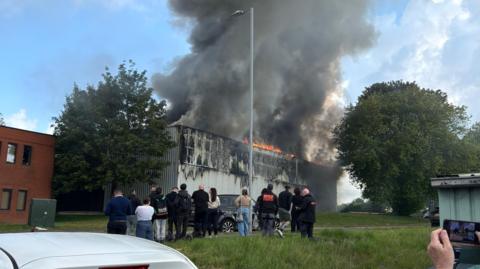 A crowd of people gather on a grassy bank as they watch a grey building with orange flames on the roof. A large plume of dark smoke is rising into the blue sky.