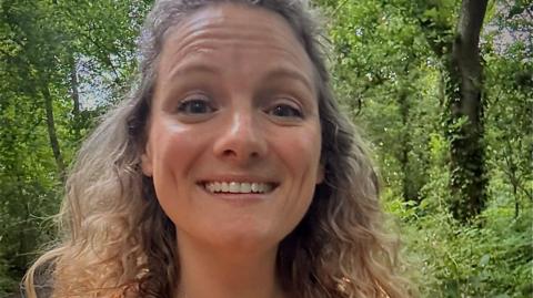 Zoe smiles at the camera in a selfie. She has light brown, curly hair and there are trees and woodland visible behind her. 