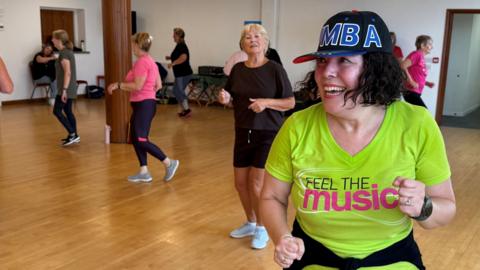 A woman with dark hair in a green top and wearing a baseball cap with the word "Zumba" on the front. She is leading a Zumba dance class in a fitness studio with some of the participants in the background.