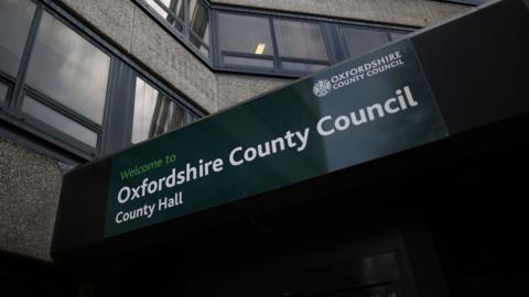 The exterior of a building as viewed from the ground, showing concrete walls and large windows. A dark green sign on the building reads "Welcome to Oxfordshire County Council, County Hall".
