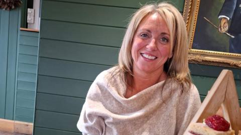 A woman with long blonde hair and wearing a grey top, smiles as she sits at a table in a restaurant.