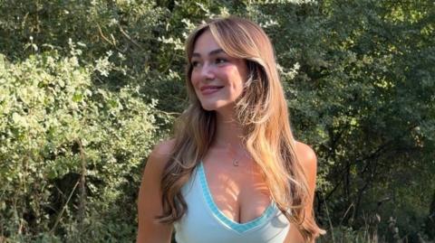 A woman with long brown hair smiling, her head is titled to the side and she is wearing a blue v-neck strapless top, she is standing in front of a woodland area.