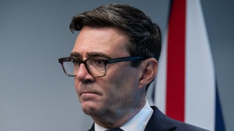 A head and shoulders shot, side on, of Prime Minister Andy Burnham. He has thick, shortish black hair, wide but neat eyebrows and dark-rimmed glasses. He is looking into the middle distance with his mouth tight shut. He wears a dark suit jacket and white shirt with a tie. Behind him is a grey wall with a union jack partially visible.