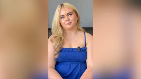 A girl with blonde hair is sitting with her head tilted to one side. She is wearing a blue summer dress.