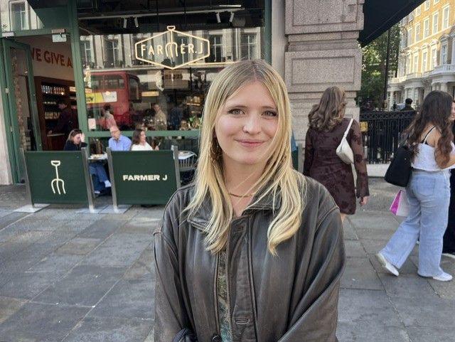 Katy, who has blonde hair and is wearing a dark jacket, smiles at the camera in a central London street.
