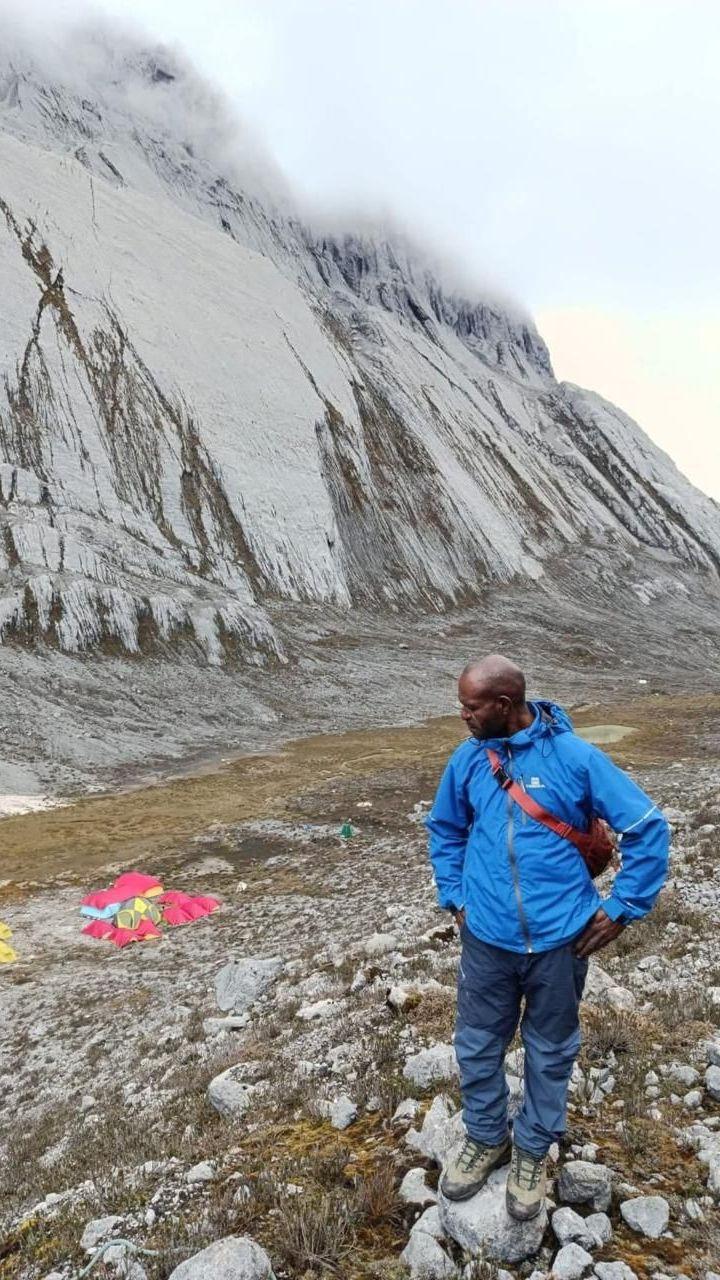 Justinus, a man from the Moni tribe, stands in front of the mountain. He is wearing a blue jacket and dark blue pants, with a red sling bag over his shoulder