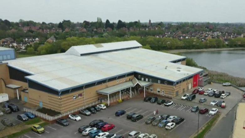 A large brown building with a white roof and covered entrance. There is a large car park in front of it and another building to the right.