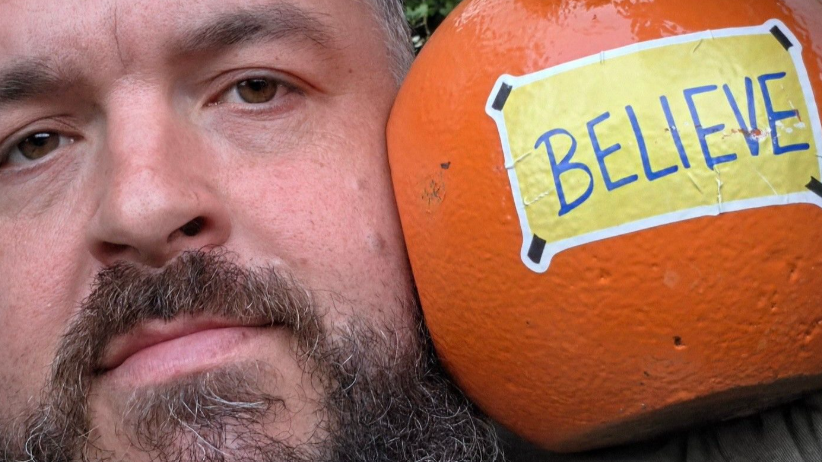 A man with a brown beard and a large orange metal ball beside his head with a yellow note on it with the word Believe in black lettering