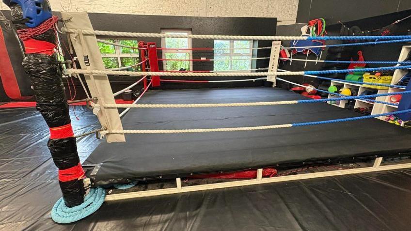 A boxing ring that looks like it has seen better days, with black bin bags wrapped around the poles. It is in a gym with black padded floors, and there are some windows behind it which look out into the forest.