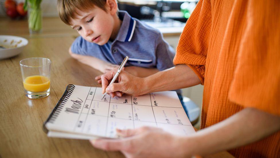 A woman holds a calendar and is writing things down, while a small child peers at the calendar