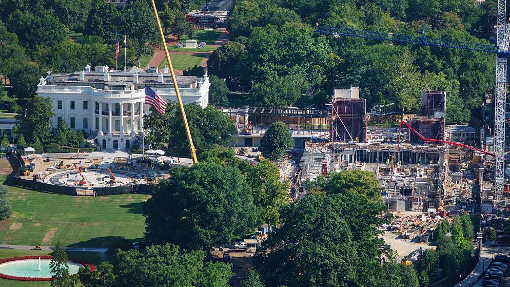 An aerial image of the White House, a large white building with tall classic columns. To the right of the building there's a large construction site, with scaffolding and cranes building a new structure. 