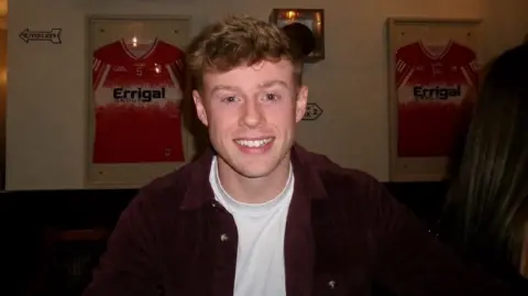 Luke McCosker has wavy reddish/brown hair and is smiling with an open burgundy coloured shirt and white t-shirt. He seem to sitting in a pub 