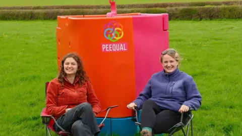 Amber Probyn and Hazel McShane sit on fold out chairs in a field in front of their product. The product is made out of plastic and coloured orange and pink, with a blue base. The words PEEQUAL is also written across the product with a logo comprised of six circles interlinking.