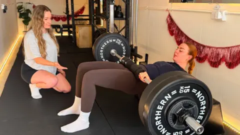 Antonia Duffin Antonia, on the left, wearing a grey t-shirt and black shorts, is talking to a young female client while she is doing hip thrusts using heavy weights on a bar which is lying across her hips