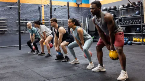 Getty Images People in a gym are lined up using kettle bells, swinging them between their legs with feet apart - behind them are shelves of weights including dumb bells and bar bells. The walls are painted grey. The floor mats are also grey.