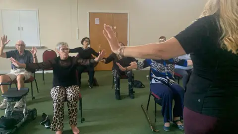 A group of people who are seated listen to music and follow choreographed movement