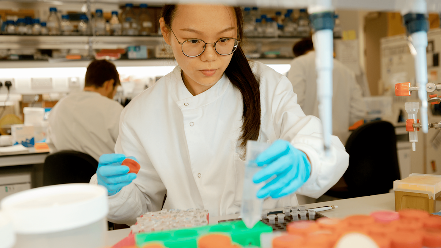 Scientist in a lab coat and blue gloves working with a pipette and sample tubes.