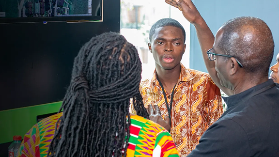 Three people engage in a discussion near a digital screen, with one man speaking.