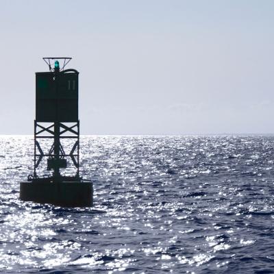 A green navigational buoy with a small light is floating in the open sea under bright sunlight and a clear sky.